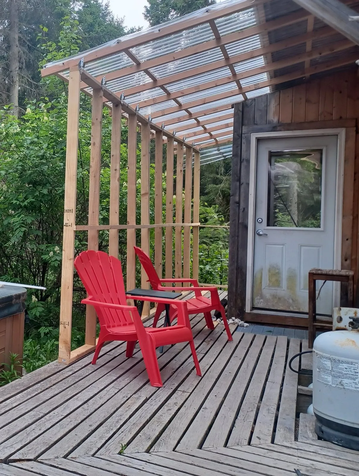 Cobertura transparente ondulada sobre um deck de madeira com cadeiras vermelhas, apoiada em estrutura de madeira junto à porta da casa